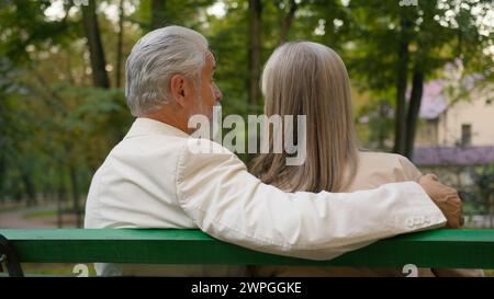 Back view cute positive 70s elderly couple retired happy family relaxing outdoors city nature park bench. Married spouse Caucasian man woman hugging Stock Photo