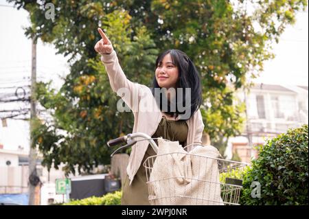 A happy young Asian female traveller in a cute dress is pointing her finger at somewhere, exploring the city with a bicycle on a bright day. Stock Photo