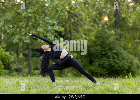 An active young woman performs a yoga pose outdoors, surrounded by vibrant green trees and the glow of the sunset. Stock Photo