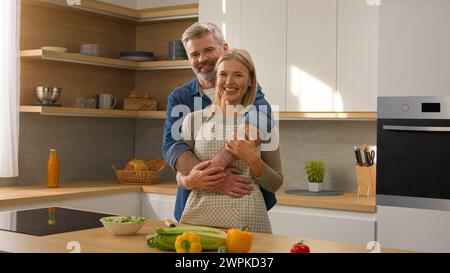 Loving happy family carefree couple adults middle-aged spouses senior man and mature woman homeowners cuddling hugging at kitchen smiling after Stock Photo