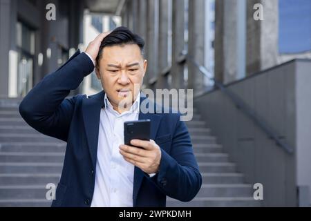 A confused Asian businessman reads a text on his phone, frowning with hand on head, standing on stairs outside. Stock Photo