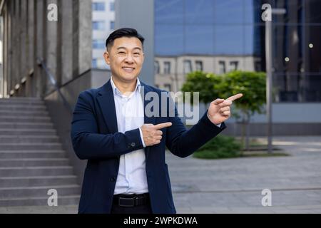 Smiling Asian businessman in a suit pointing with both hands to the side, standing on a city street with buildings in the background. Stock Photo