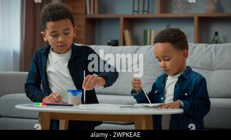 Two African American children kids school boys ethnic little friends siblings brothers drawing picture with colored paints painting in living room Stock Photo