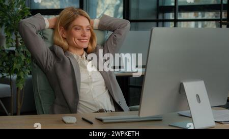 Smiling calm relaxed 40s business woman finished computer work stretching sitting at desk in modern office. Happy middle-aged businesswoman feeling Stock Photo