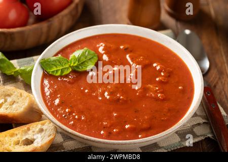 Bowl of tomato soup garnished with basil leaves and toasted baguette slices, fresh tomatoes in background on wooden table Stock Photo