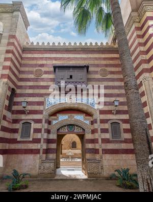 Striking red and white striped arched entrance leads into a Mamluk era style building, showcasing intricate architectural details surrounded by lush palm trees Stock Photo