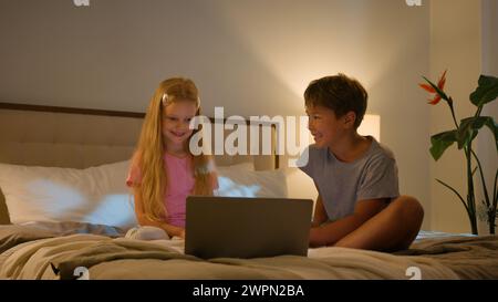 Laughing Caucasian happy children brother sister kids girl boy looking laptop screen in bedroom on bed evening watching cartoon video online using Stock Photo