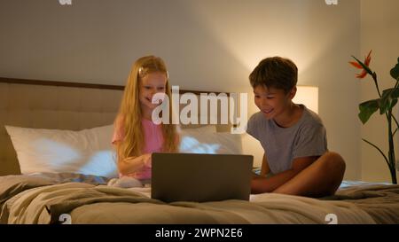 Laughing Caucasian happy children brother sister kids girl boy looking laptop screen in bedroom on bed evening watching cartoon video online using Stock Photo