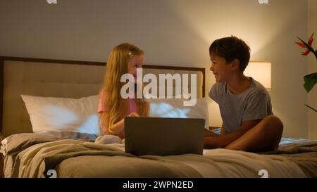 Laughing Caucasian happy children brother sister kids girl boy looking laptop screen in bedroom on bed evening watching cartoon video online using Stock Photo