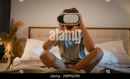 Surprised amazed Caucasian child kid boy playing video game using innovative digital VR headset on bed home play online 3d AR technology in metaverse Stock Photo