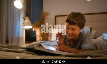 Surprised amazed smiling funny Caucasian child kid boy schoolboy reading interesting book evening in bedroom using flashlight lying on bed home Stock Photo