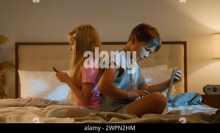 Modern Caucasian children kids boy girl brother sister siblings playing online video game together using different digital gadgets on bed at home Stock Photo