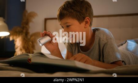 Surprised amazed smiling funny Caucasian child kid boy schoolboy reading interesting book evening in bedroom using flashlight lying on bed home Stock Photo