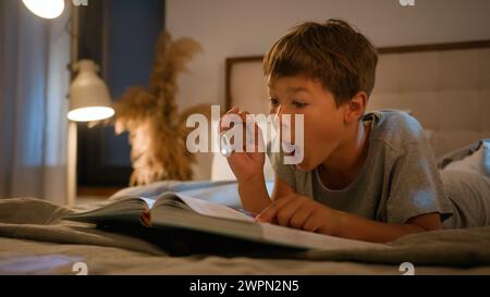 Surprised amazed smiling funny Caucasian child kid boy schoolboy reading interesting book evening in bedroom using flashlight lying on bed home Stock Photo