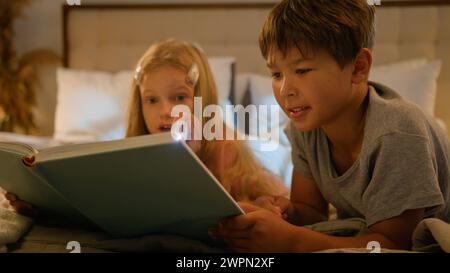 Caucasian children kids boy girl older brother younger sister lying on bed at home evening reading interesting book using flashlight read fairytale Stock Photo