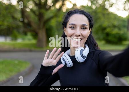 Cheerful young woman with headphones enjoying an outdoor fitness routine while taking a selfie in the park. Stock Photo