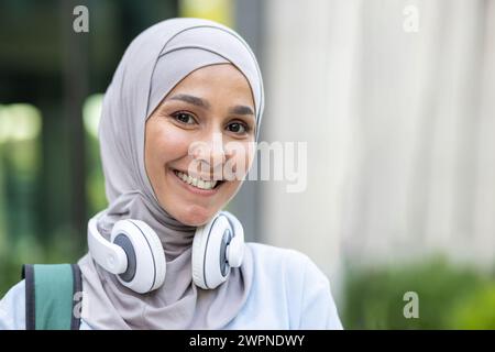 Portrait of a smiling young Muslim woman wearing a hijab and headphones around her neck, expressing happiness in an urban setting. Stock Photo