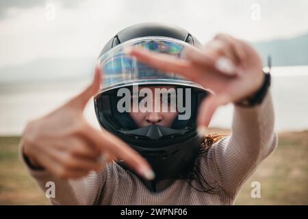 A creative shot of a Latina woman framing the camera with her hands while wearing a motorcycle helmet, with a lake backdrop Stock Photo