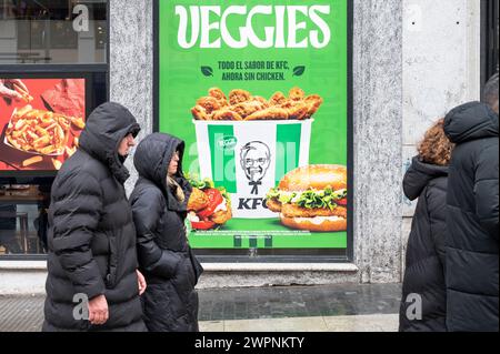 People walk past the American fast-food chicken restaurant chain, Kentucky Fried Chicken (KFC) street commercial advertisement featuring a new vegetarian meal named Veggies available at their fast-food chains in Spain. Stock Photo