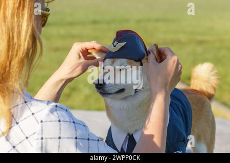 A woman dresses her own Shiba Inu dog in a pilot suit at the airport. Funny cosplay with dressing up a dog in human clothes. Stock Photo