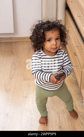 A mixed race curious toddler holds a TV remote, symbolizing the early interaction with digital devices in modern childhood, and development. High qual Stock Photo