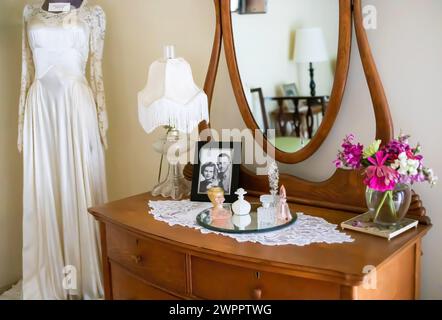 Antique dresser and mirror with an assortment of perfume bottles, wedding picture, flowers, lamp, and a wedding dress in the corner in a bride's room. Stock Photo