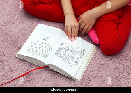 A Young girl child sitting on a soft pink surface, reading an open book written in Polish language. Relaxed and focused, with the book open on an illu Stock Photo