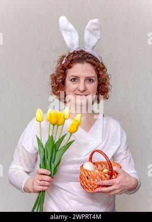 A woman with bunny ears on her head holds a wicker basket with Easter eggs and a bouquet of yellow tulips. Stock Photo