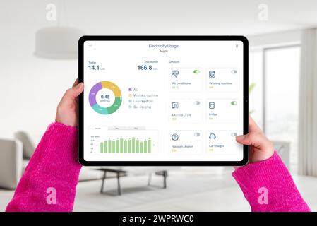 Hands hold tablet in living room with electricity usage app, monitoring consumption and controlling home devices. Concept of smart energy management Stock Photo