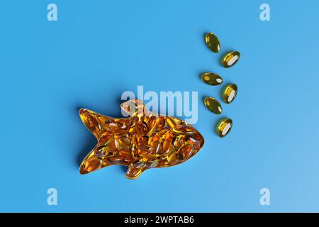 Fish shaped tin box filled with orange elliptical fish oil pills and some capsules forming bubbles on blue background. Omega-3 fatty acid supplements Stock Photo