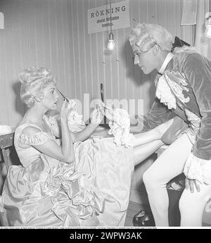 As if in the 18th century. A man and a woman dressed in stage clothes before a performance backstage. She gets help holding the small make-up mirror when she has to paint her eyebrows. Both are wearing their historical clothes that suggest a historical scene. The lodge is simple with only light bulbs as lighting and the no smoking sign on the wall. Probably an open-air theater in Sweden in 1948. Kristoffersson ref AL10-8 Stock Photo