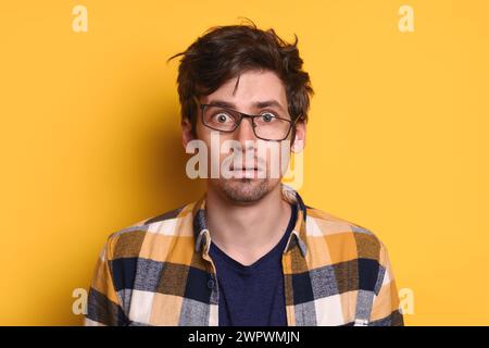 Shocked puzzled caucasian man in eyeglasses with wild hair opened mouth posing on yellow studio wall, has surprised expression on face. Overworked freelancer feel exhausted. Emotions, shock concept Stock Photo