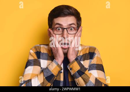 Portrait of shocked young man, stares at camera with stupefied expression, opens mouth, says wow, keeps hand on cheeks, isolated on ver yellow background. People, reaction and emotions concept Stock Photo
