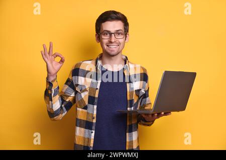 Portrait of positive smart confident cool man in eyeglasses holding laptop, making okay hand gesture isolated on yellow background, finding new clients on social media, online learning, technology Stock Photo