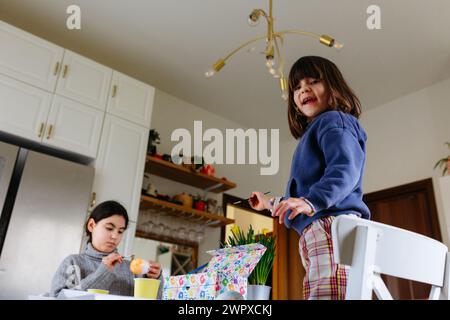 sitting girl at the table coloring the egg while smaller girl stands on chair smiling looking at camera Stock Photo
