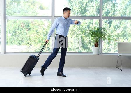 Handsome steward with suitcase looking at wristwatch in hall of airport Stock Photo