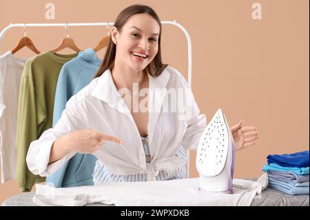 Pretty young woman pointing at modern iron on ironing board against beige background Stock Photo
