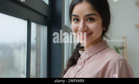 Satisfied girl dreamy Arabian woman looking through window in apartment waiting happy young female businesswoman calm lady dreaming peaceful morning Stock Photo