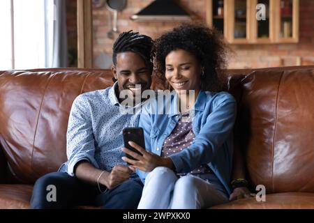 Attractive African couple sit on sofa looking at cellphone screen Stock Photo