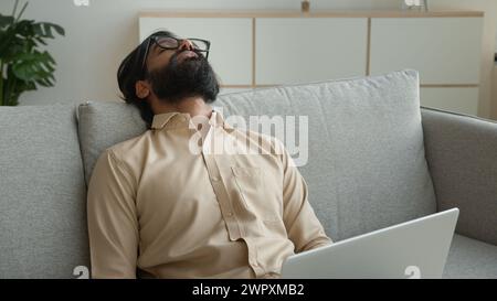Tired exhausted freelancer working with laptop at home overworked Arabian muslim man fall asleep on couch sick weary sleeping Indian businessman lean Stock Photo