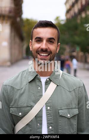Portrait young bearded handsome man stands looking at camera smiling on street in European city. Stock Photo