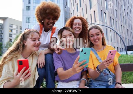 Multiracial group of young cheerful women gathering together outdoors in the city, using cellphones. Stock Photo