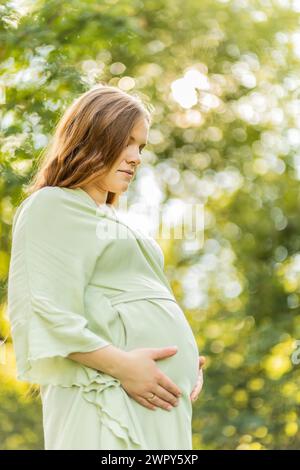 Young pregnant woman holds big belly, green trees,sky on background. Brown long haired female wear dress. Magic happy pregnancy. Child delivery Stock Photo