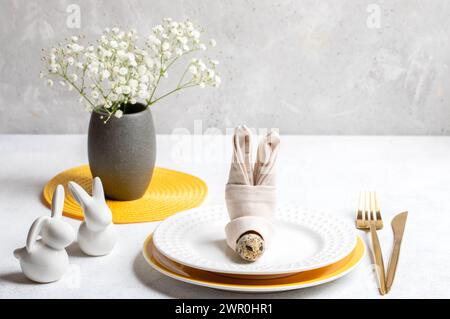 Festive light elegant Easter table setting. Napkin as bunny muzzle, white and yellow plates, golden cutlery, small bunnies, white Gypsophila flowers o Stock Photo