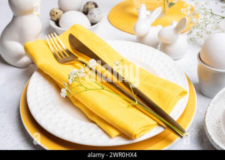 Fragment of holiday Easter table. Serving in white with yellow colors. Golden cutlery on plates surrounded by ceramic bunnies, natural colors eggs, de Stock Photo
