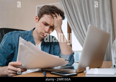 Unpappy young man sitting in home office and looking thoughtful on papers, thinking of home budget, using laptop for paying bills online, lost job due to crisis or quarantine. Money problem, bad news Stock Photo