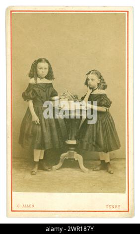 Original Victorian Carte de Visite (visiting card or CDV) of two serious looking young Welsh sisters, Victorian girls, Victorian children ,wearing fashionable crinoline style dresses, puff sleeves and boots, with long crimped hair held with a bow, pendants and holding toys possibly. From the photographic studio of C. Allen, Tenby in Wales. Circa 1864 from puff sleeves, hairstyle, dress. Stock Photo