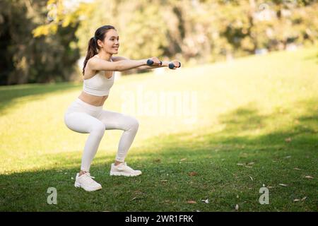 Young athletic woman in white sportswear doing squats with dumbbells in a sunny green park Stock Photo