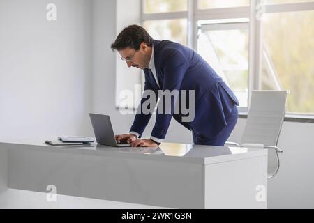 A professional man in a blue suit is intently typing on a laptop on a white desk Stock Photo