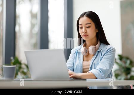 Focused young asian woman with laptop and headphones in modern office Stock Photo